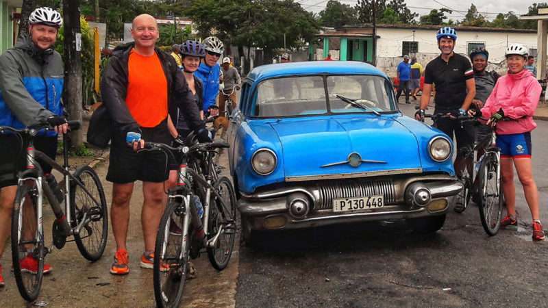 A group of cyclists pose in front of a vintage car in Cuba