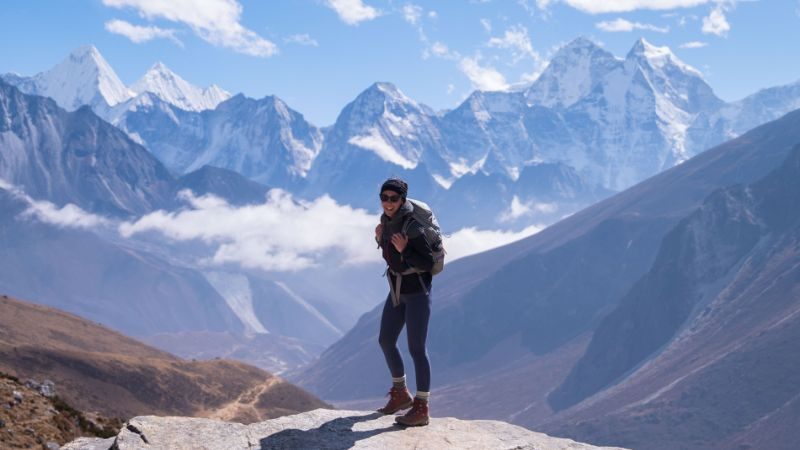 Person standing with the peaks of the Himalayas in the background wearing a backpack and similing